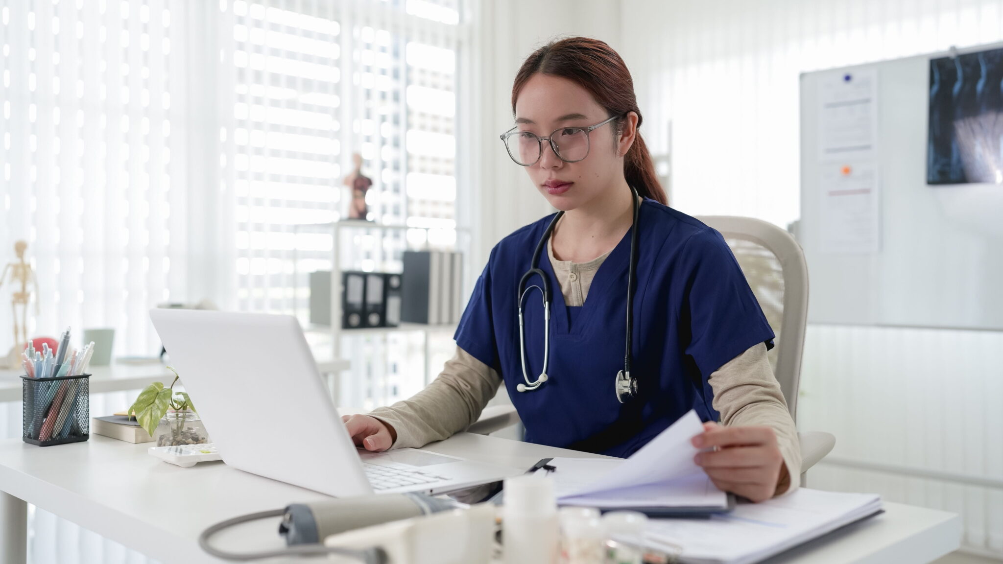 A doctor reviewing documents beside a laptop in a clinic.